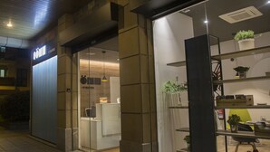 A hotel entrance with a modern design, featuring a glass door, a reception desk inside, and a visible staircase leading to the upper floors.