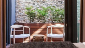Ein Balkon mit zwei weißen Stühlen und einem kleinen runden Tisch, eine Steinmauer im Hintergrund und eine Tür, die zum Zimmer führt.