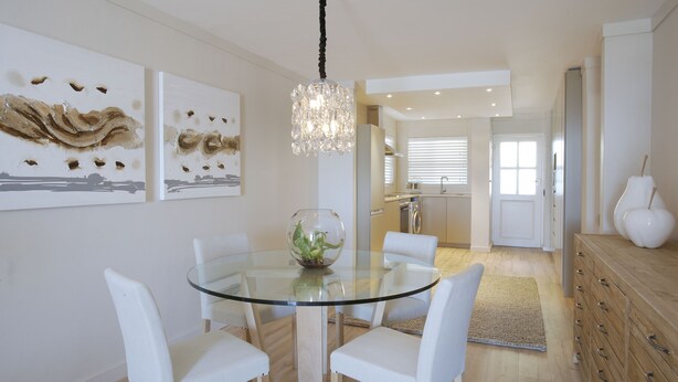 Een moderne eetruimte met een glazen tafel, witte stoelen, een kroonluchter en kunstwerken aan de muren.