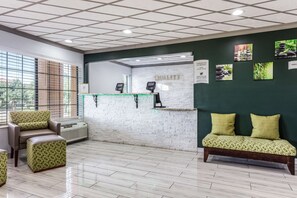 A reception area with a white brick wall, a reception desk, and a green sofa with yellow cushions. There are two chairs and a small table with a plant.