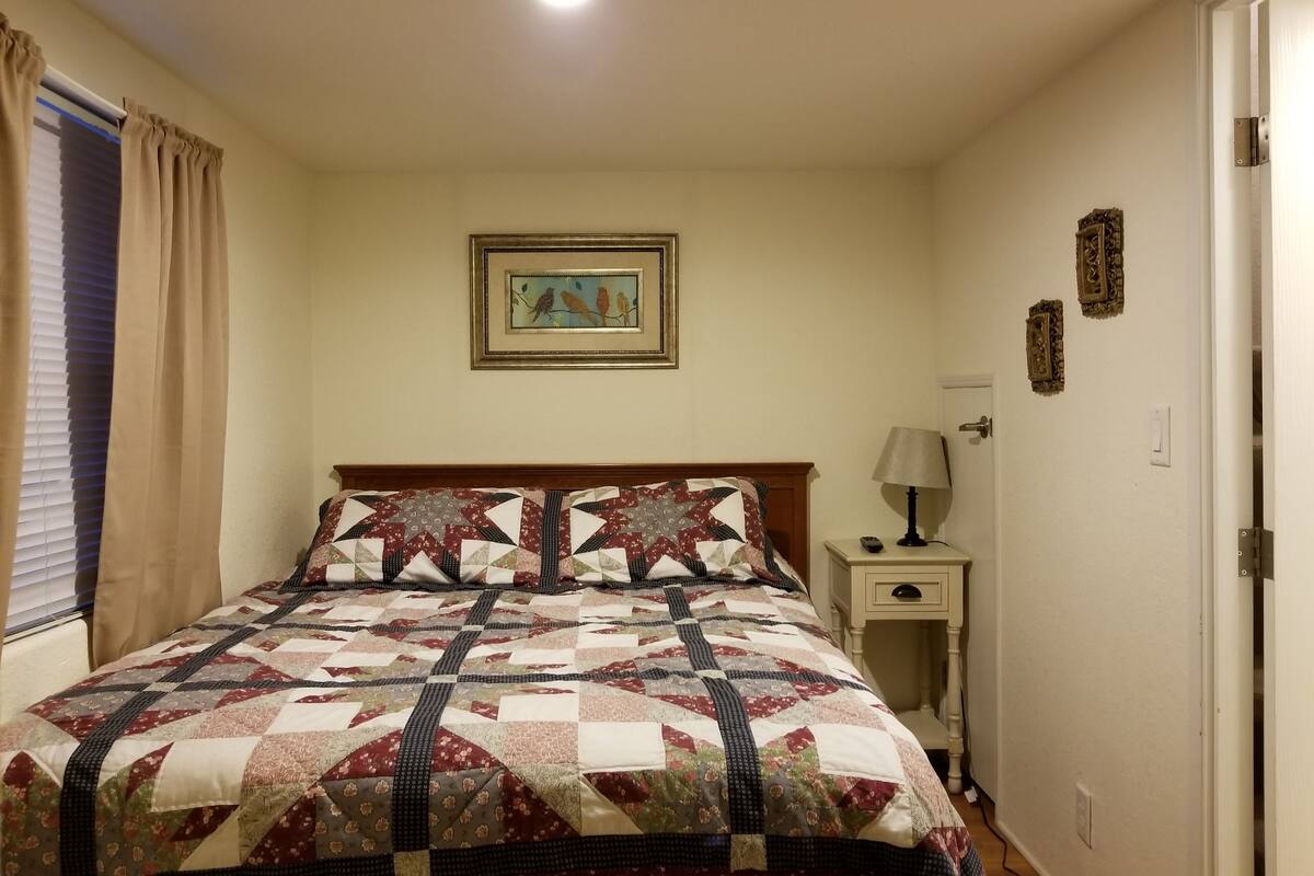 A bedroom with a large bed, a nightstand, a lamp, a framed picture on the wall, and a window with curtains.