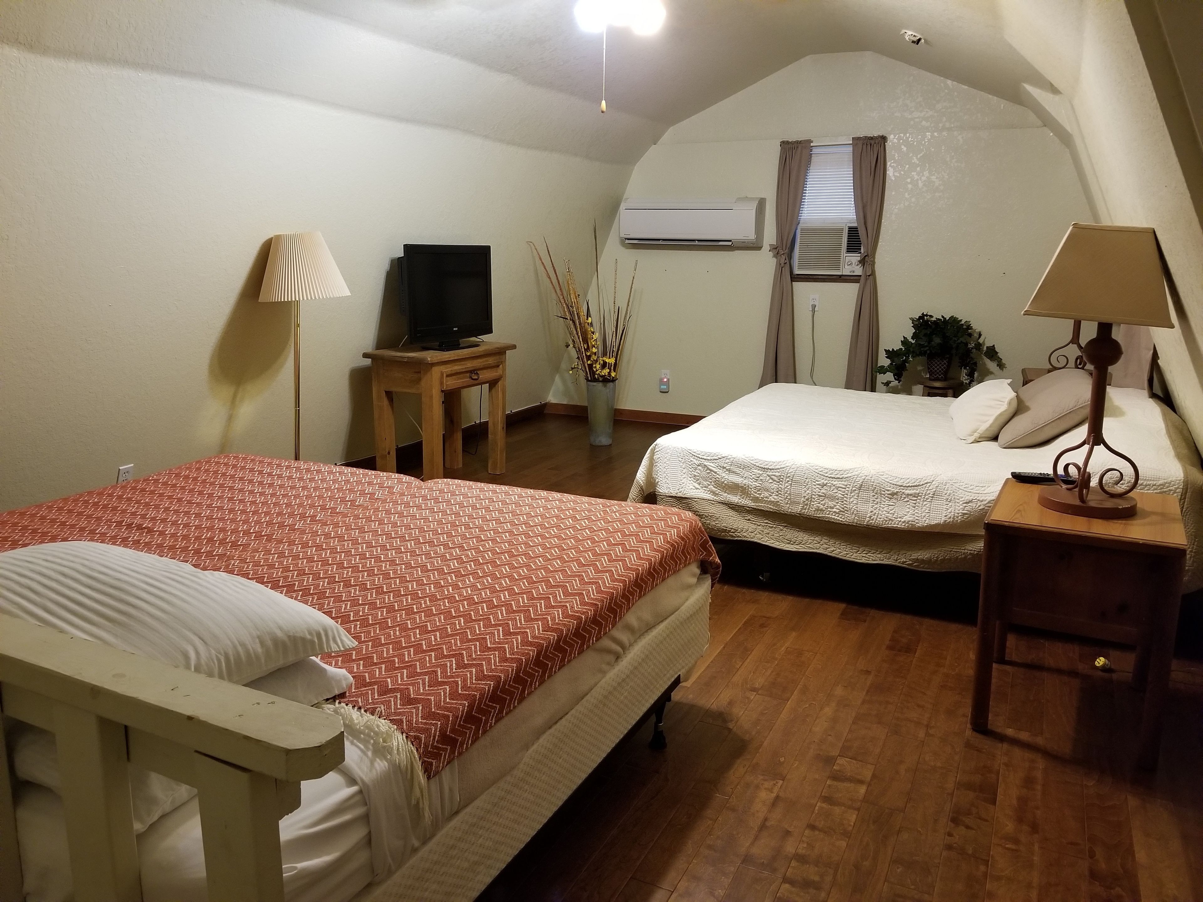 A bedroom with two beds, a TV on a wooden table, an air conditioner, and a lamp on a bedside table.