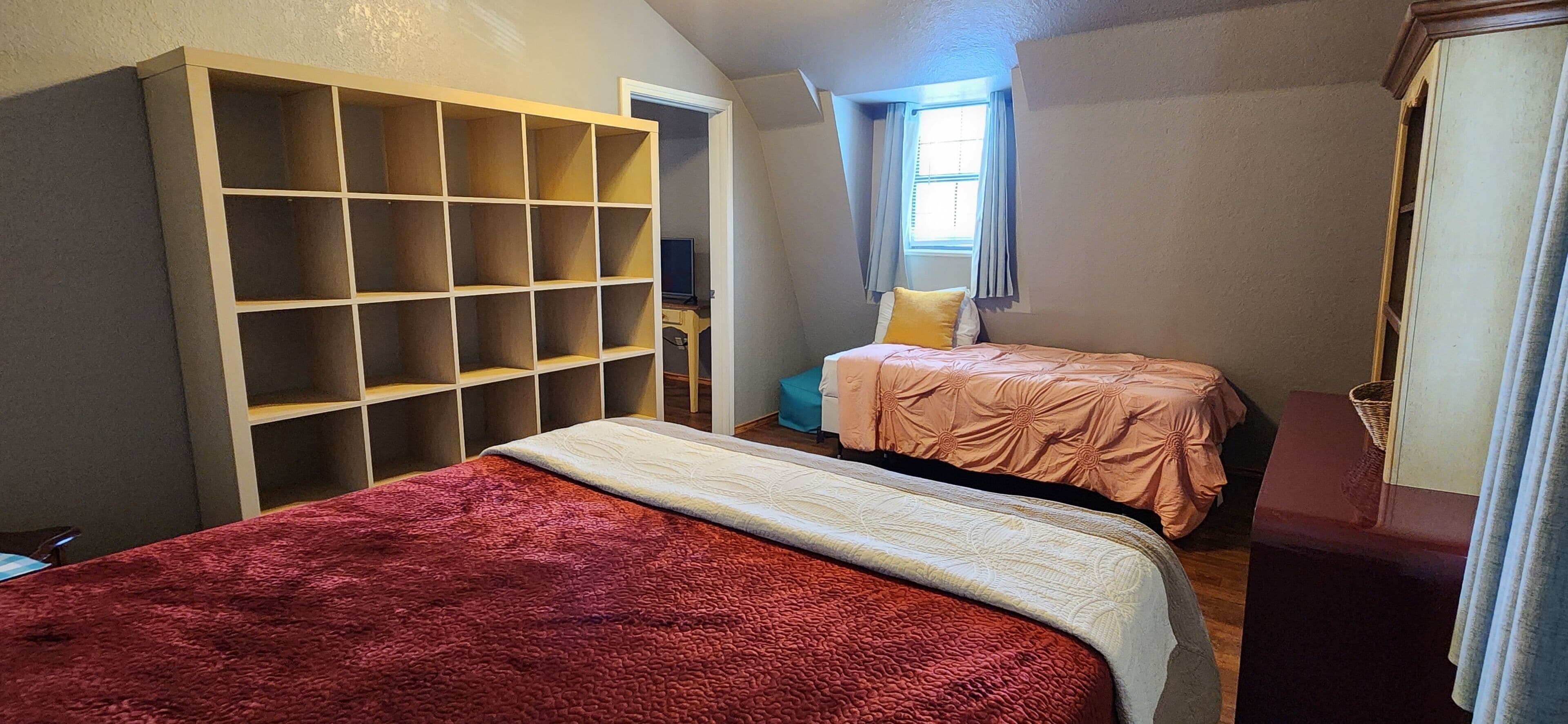 A bedroom with two beds, a large shelving unit, and a window with curtains.