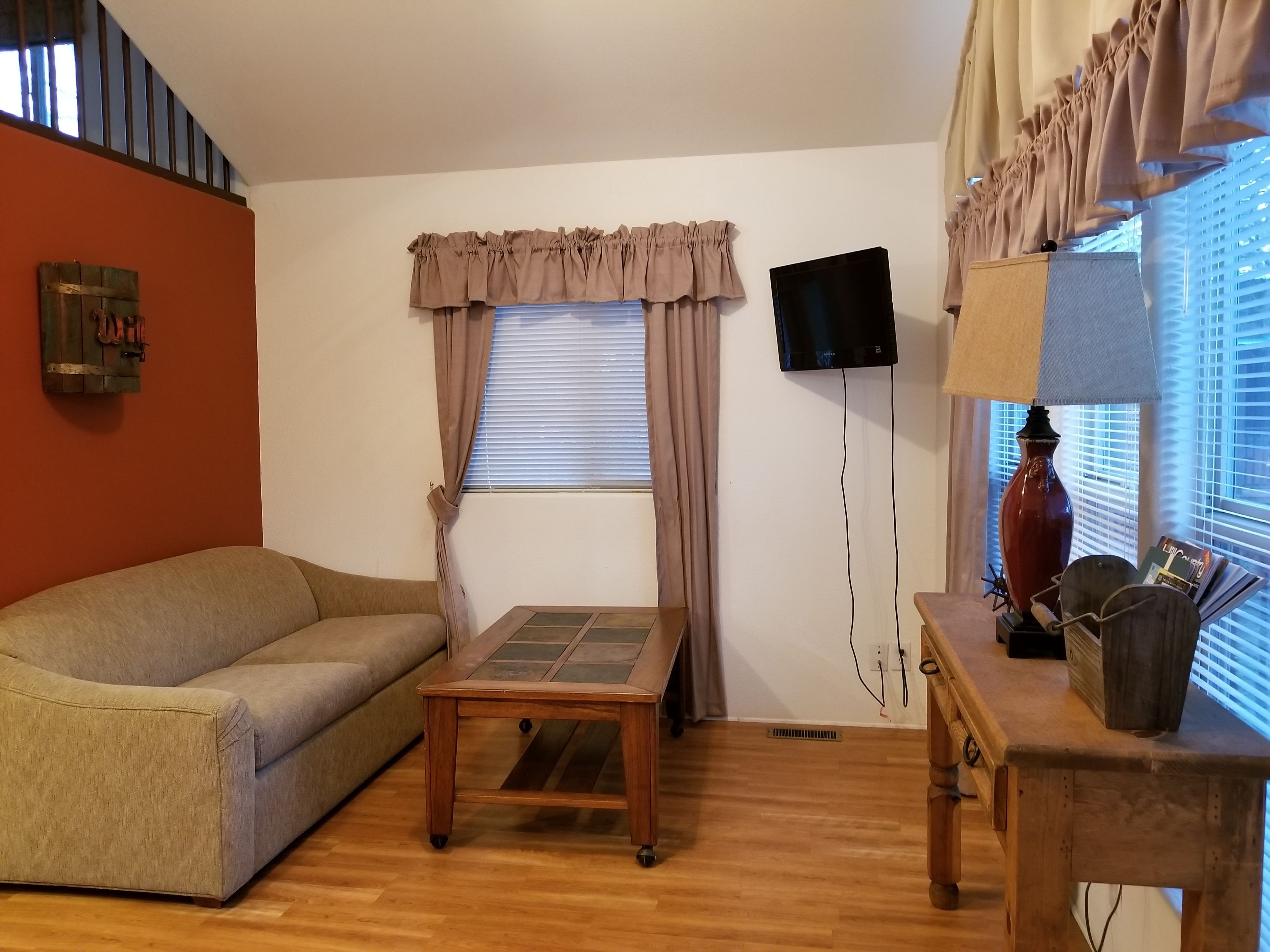 A living room with a sofa, a small table, a TV mounted on the wall, a lamp, and a window with curtains.