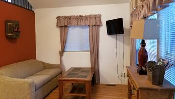 A living room with a sofa, a small table, a TV mounted on the wall, a lamp, and a window with curtains.