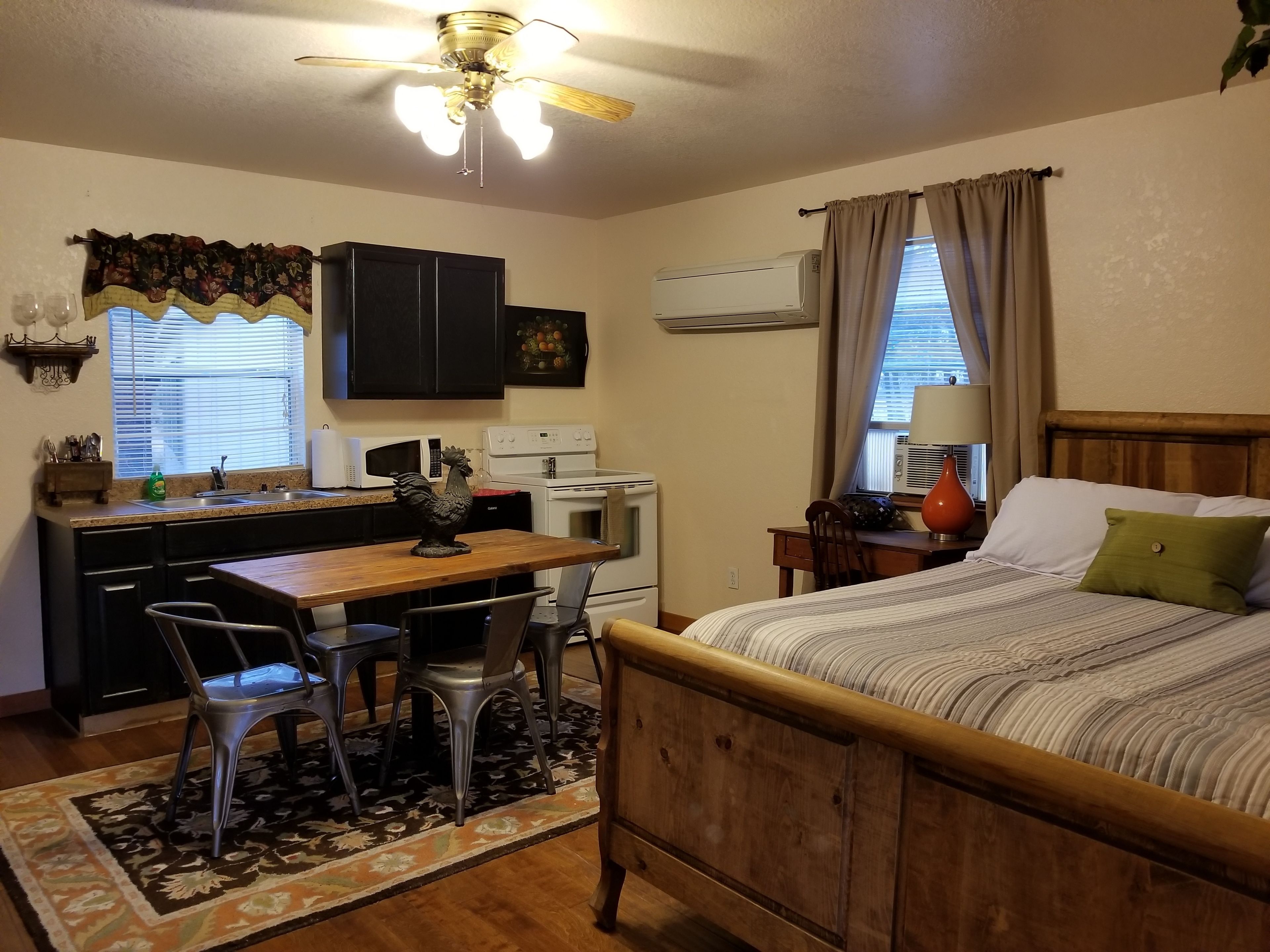 Une chambre compacte comprenant un lit en bois, un bureau avec une chaise, un petit coin cuisine et un ventilateur de plafond.