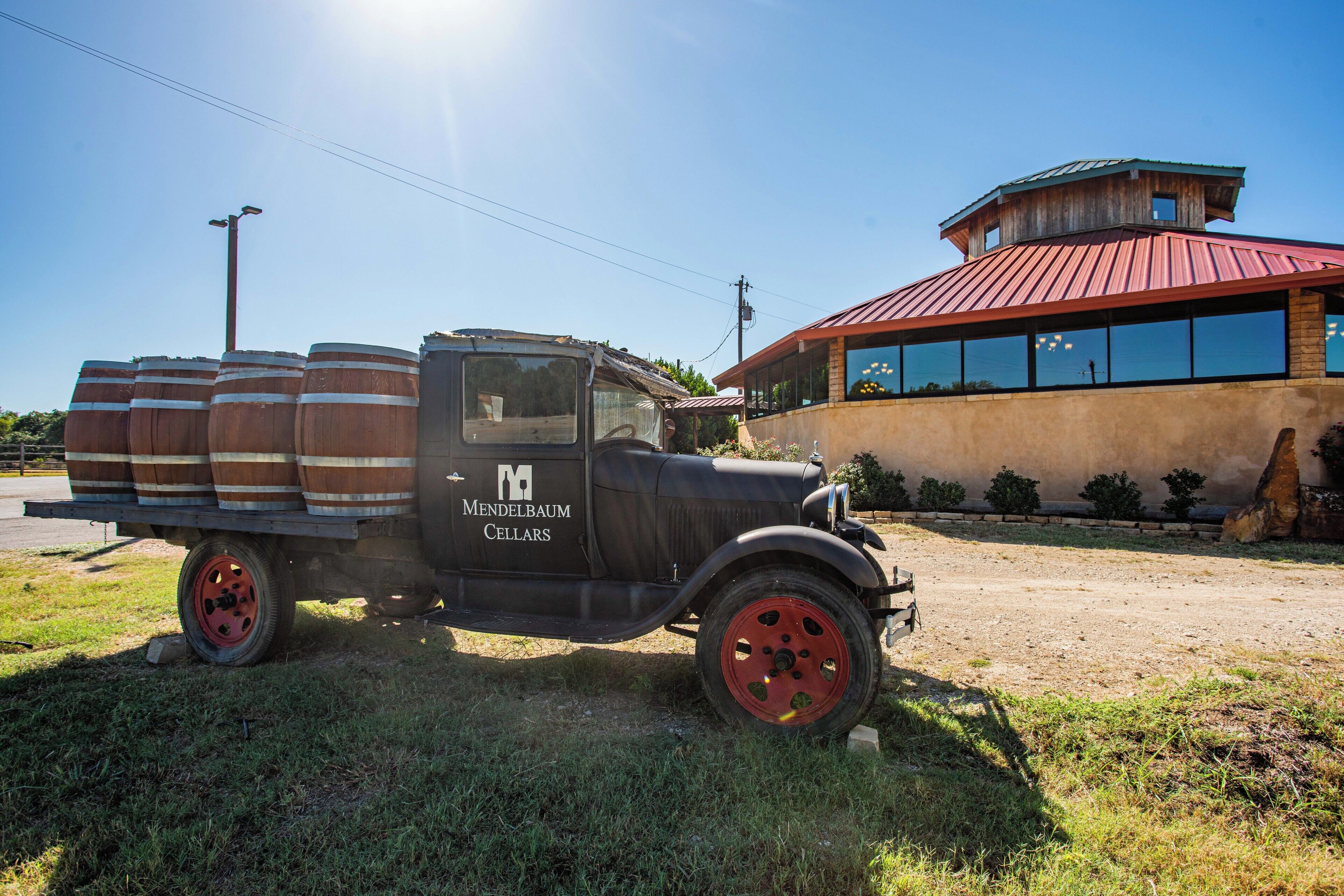 A vintage black truck with wooden barrels on the back parked on grass, in front of a building with a red roof and large windows.
