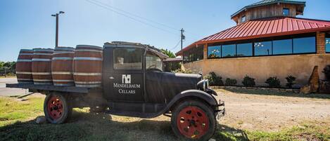 A vintage black truck with wooden barrels on the back parked on grass, in front of a building with a red roof and large windows.
