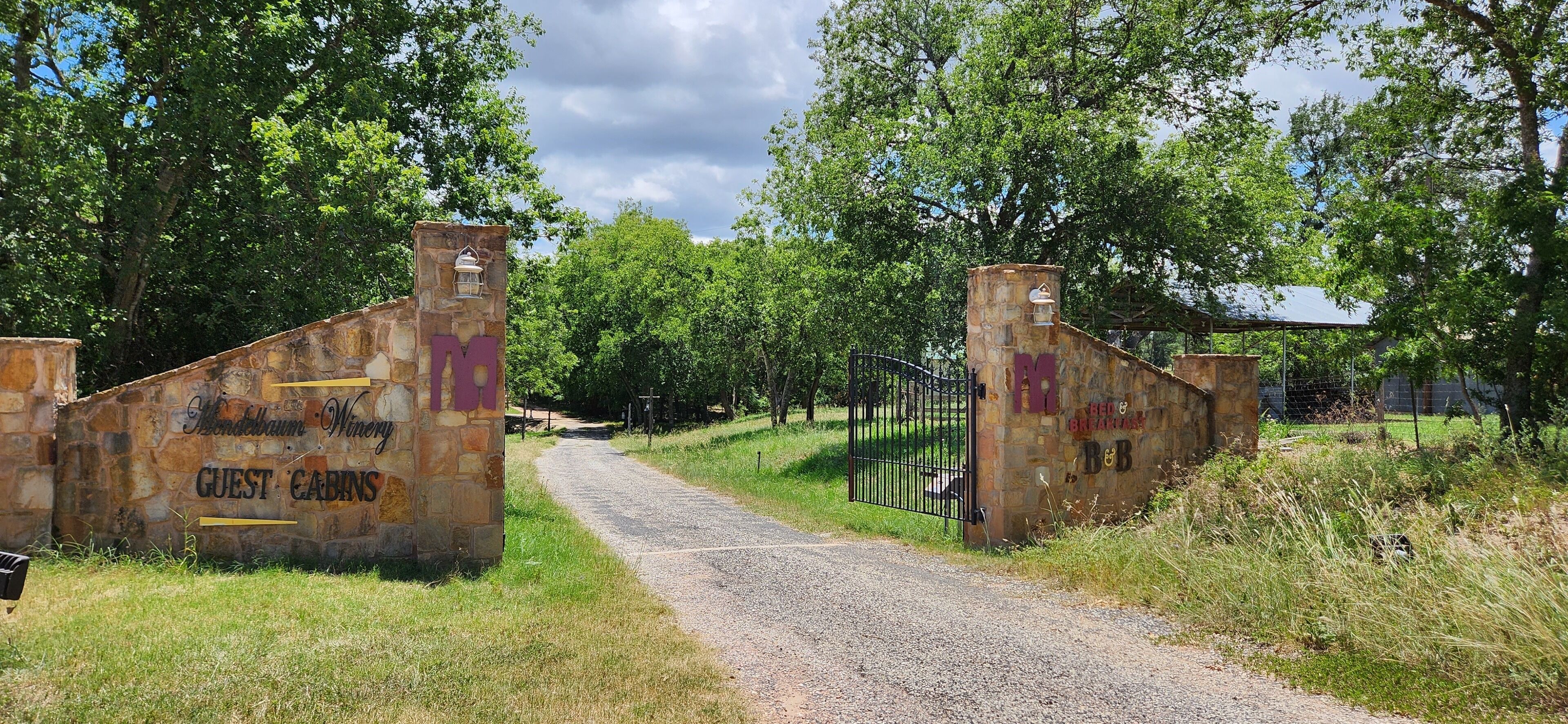 A rustic stone gate with "GUEST CABINS" and "Blondbäum Winery" signs, leading to a gravel path surrounded by trees.