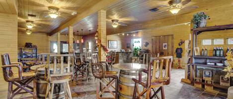 A rustic interior with wooden walls and ceiling, a bar area with bottles, and a seating area with wooden chairs and barrels.