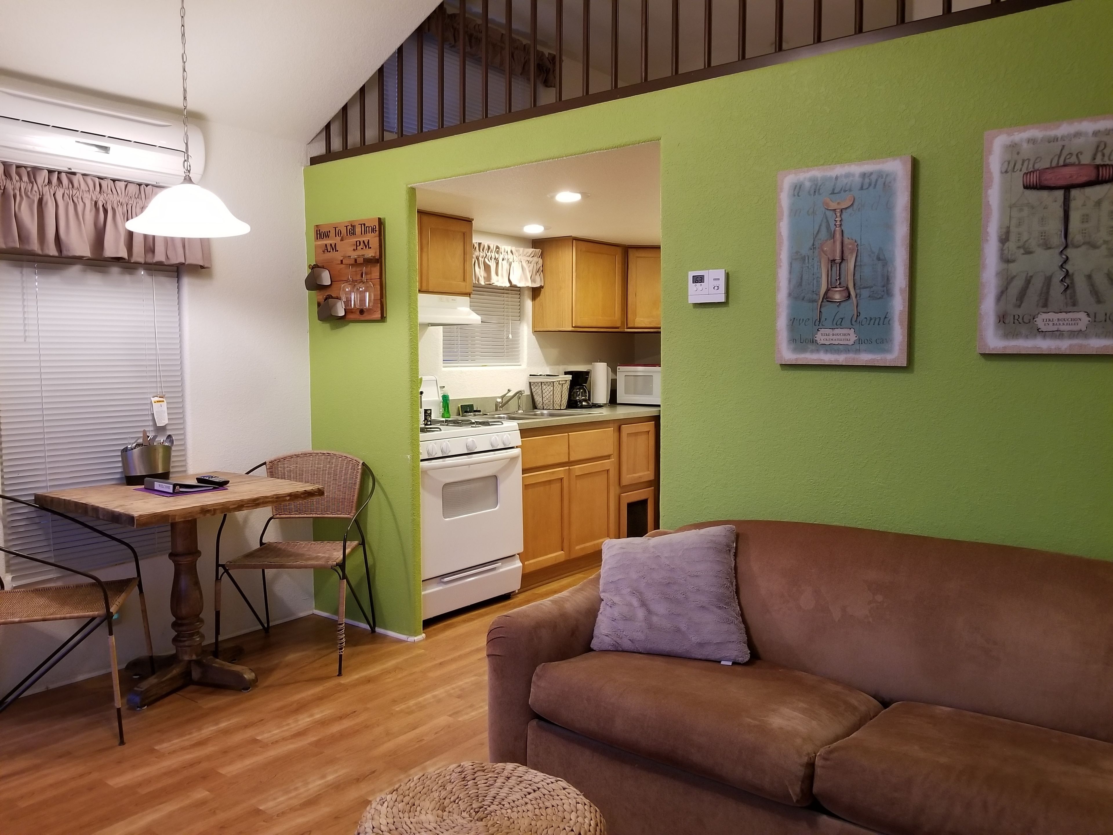 Un salon chaleureux avec un canapé marron, une table à manger avec des chaises, un coin cuisine doté de meubles en bois et un mur d’accent vert.