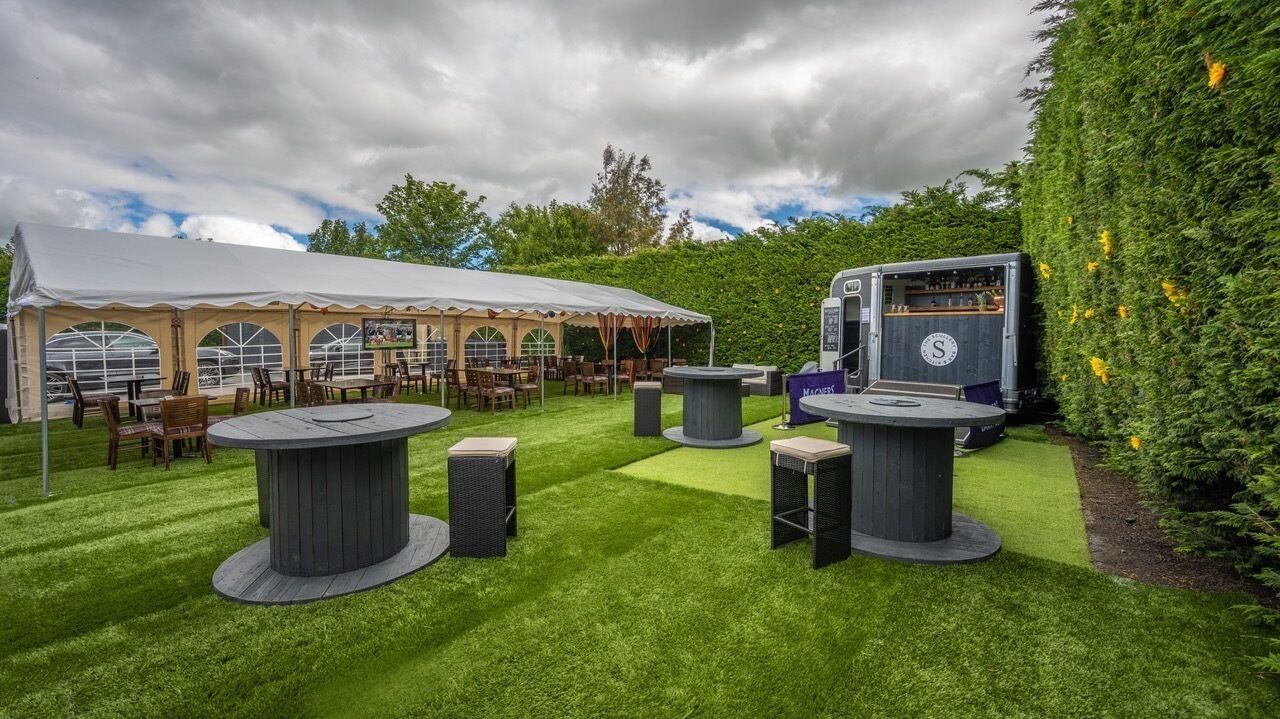 A tented outdoor area with round tables and chairs, a bar area with a counter, and a hedge on one side.
