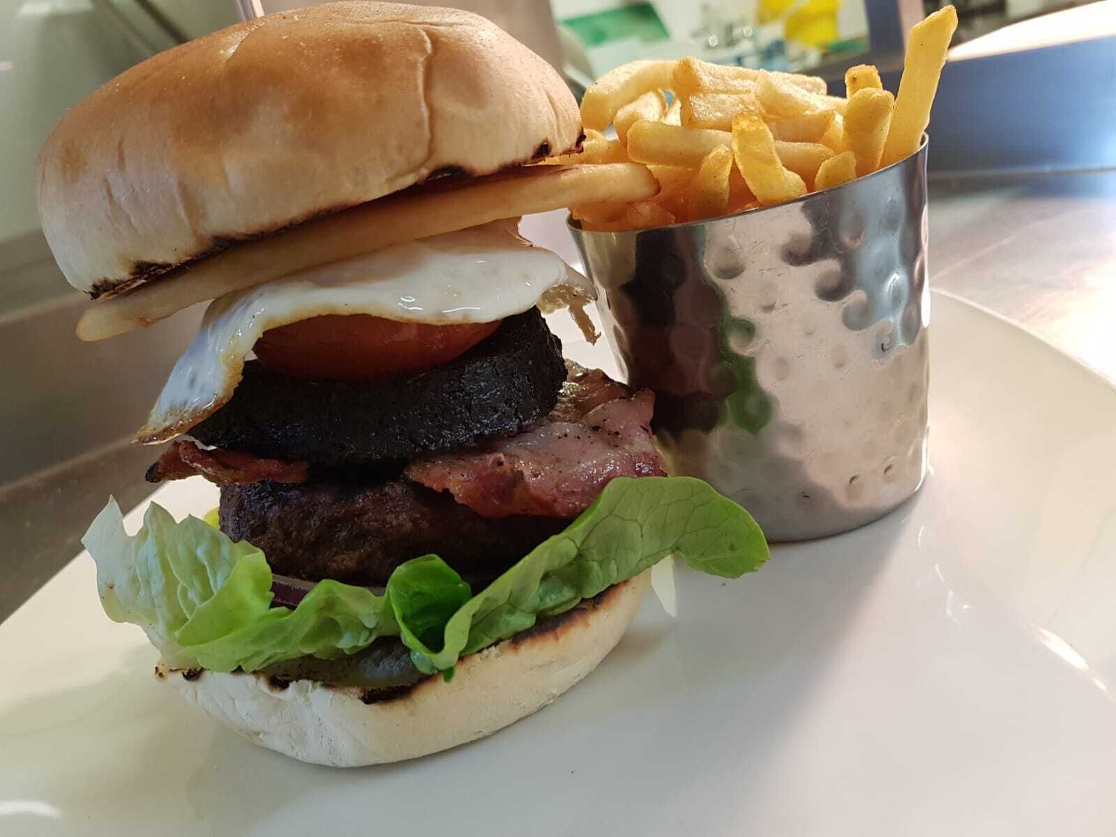 A burger with a bun, lettuce, tomato, egg, and bacon, and a cup of fries.