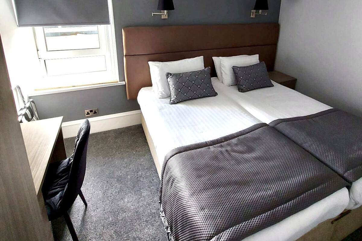 A hotel room with a bed, two bedside tables, a desk, a chair, and a window.