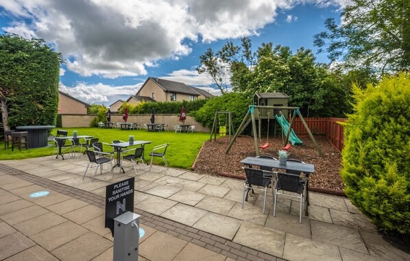 An outdoor patio area with tables and chairs, a children's play structure, and a sign instructing to sanitize hands. - The Newmachar Hotel (Aberdeen)