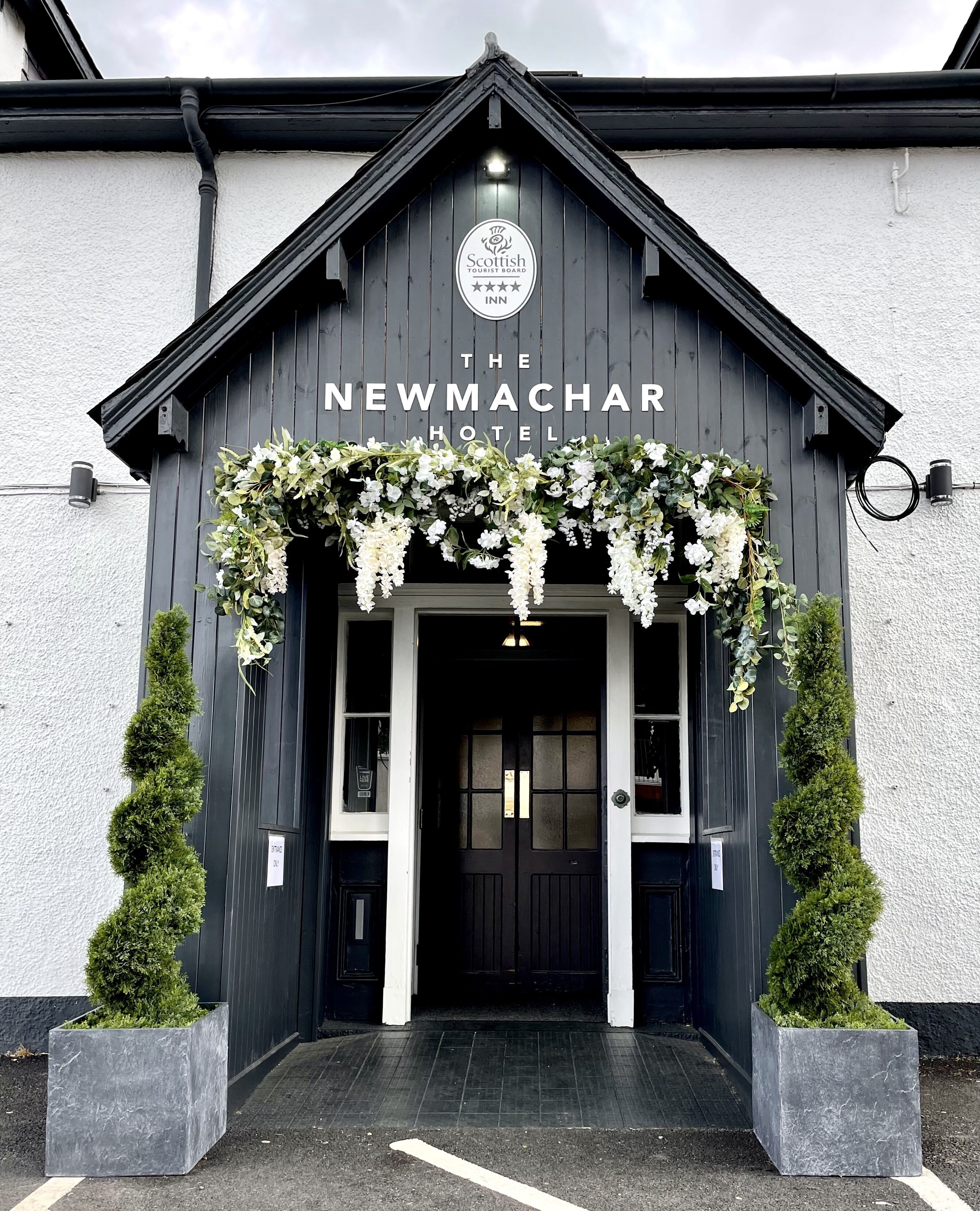 El exterior del Hotel Newmachar, con fachada oscura, un letrero que indica que es una posada del Scottish Tourist Board, y un adorno floral sobre la entrada.
