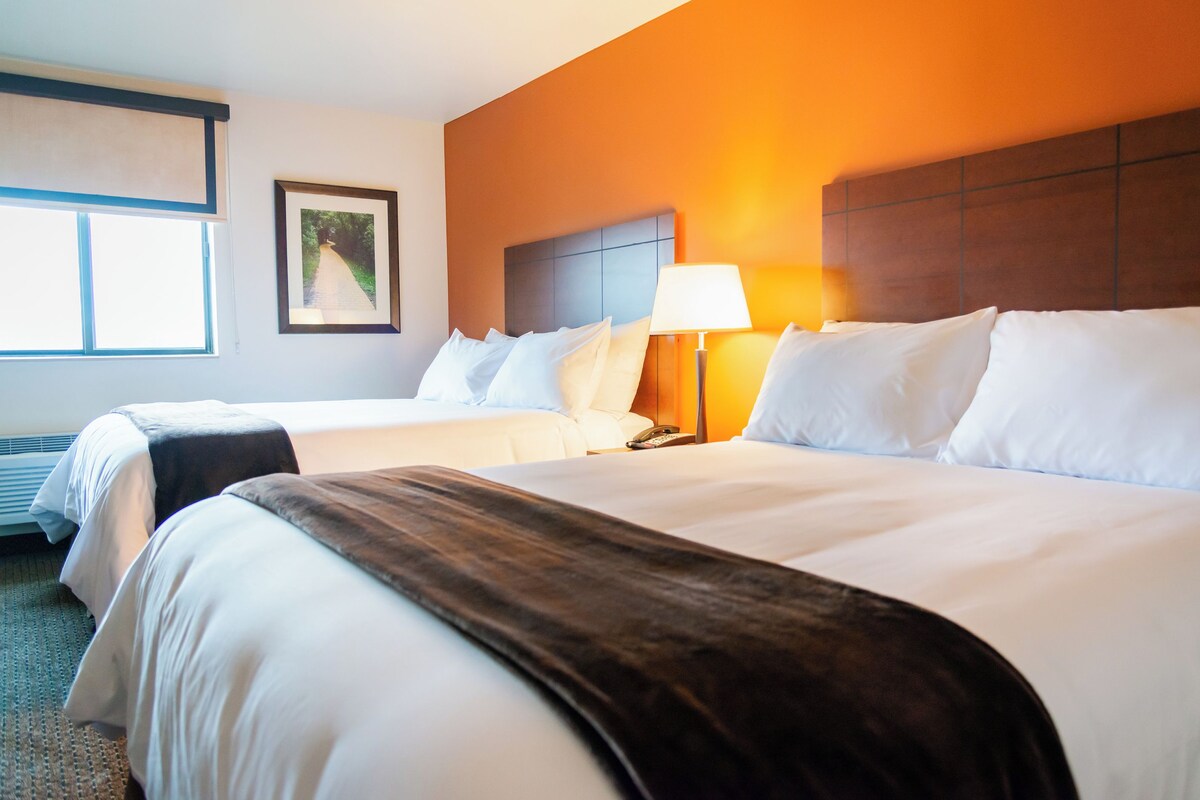 A hotel room with two beds, a nightstand, a lamp, a framed picture on the wall, and a window.