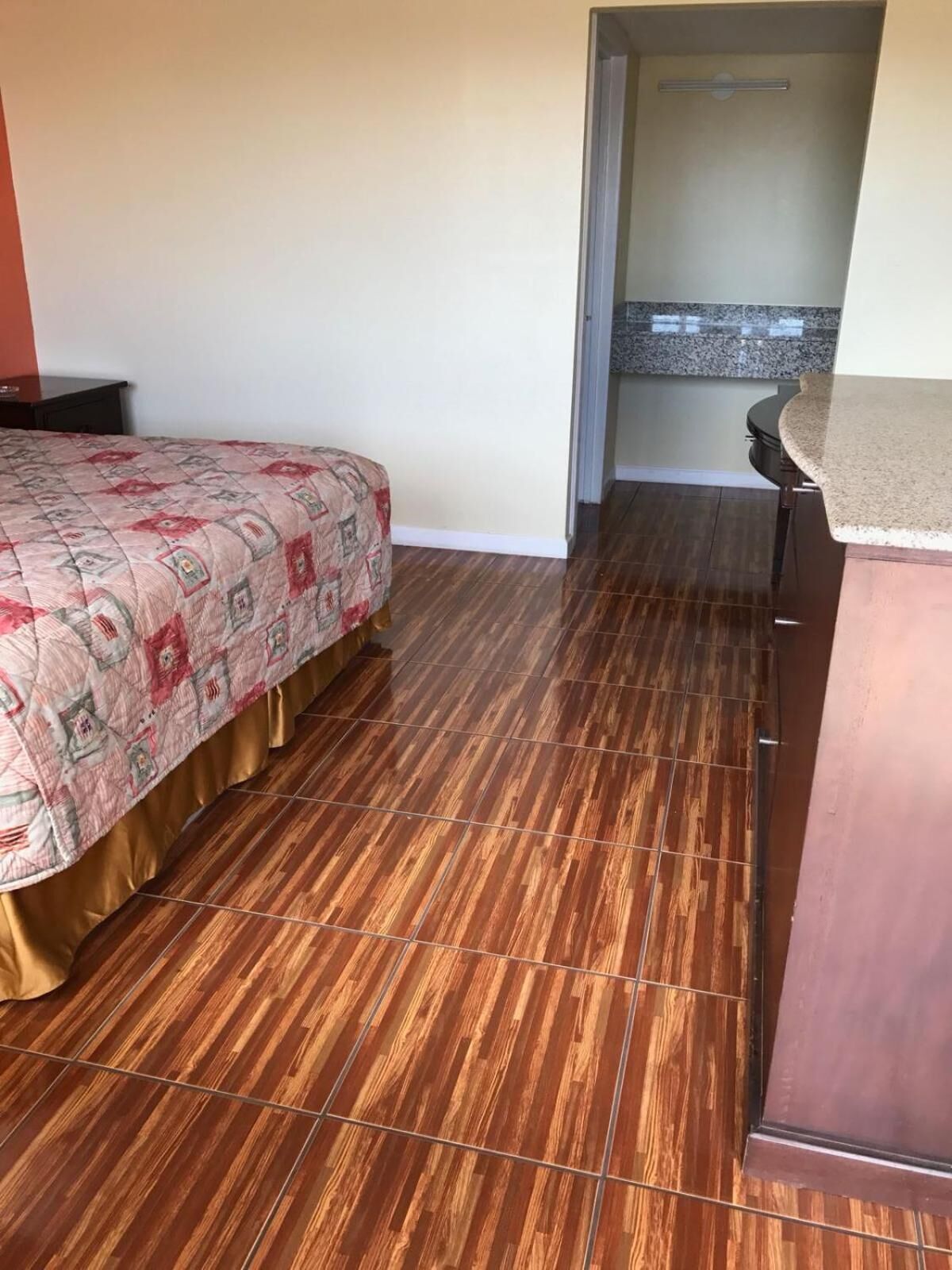 Camera d'albergo con pavimento in legno, un letto con copriletto decorato, un comò di legno e un bagno visibile attraverso una porta aperta.