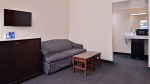 A hotel room with a flat-screen TV mounted on the wall, a patterned sofa, a small wooden table, a kitchenette with a microwave, and a trash bin. - Scottish Inns & Suites (Lubbock)