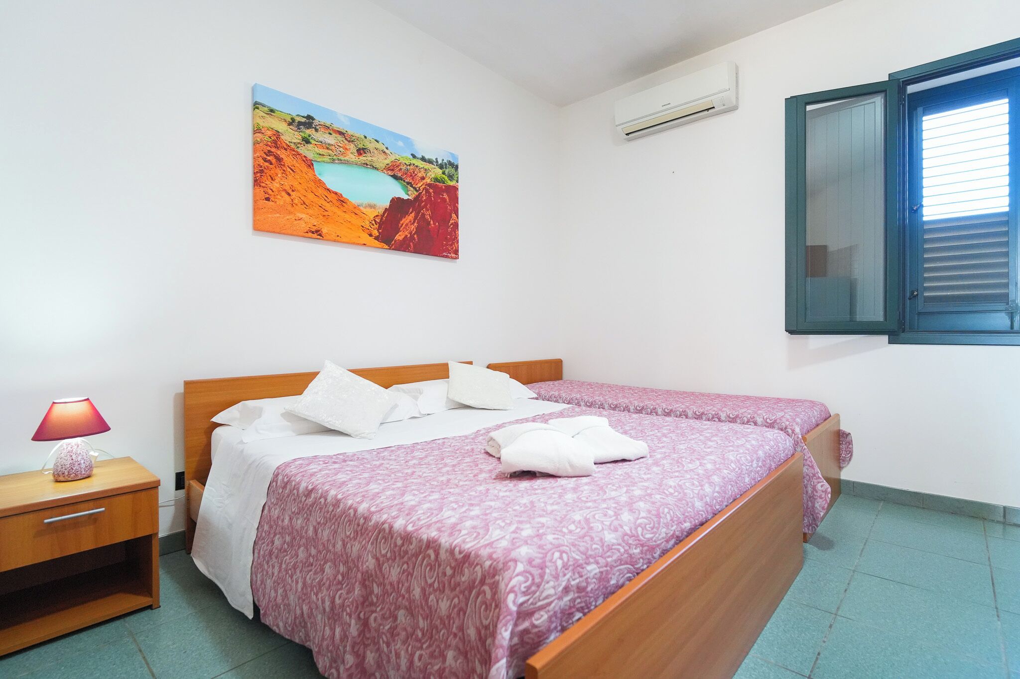 A bedroom with two beds, a nightstand, a lamp, a painting, and a window with blinds.