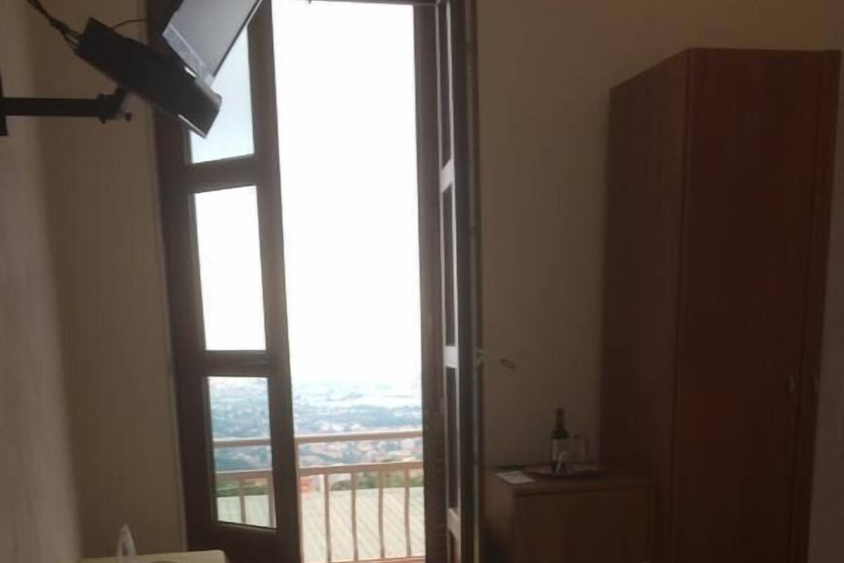 A bedroom with a bed, a wooden wardrobe, a desk with a chair, and a window with a view of the city.