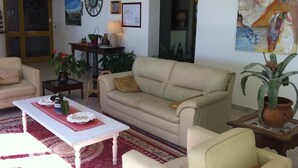A living room with a beige sofa, a white coffee table, and a red rug. There are two chairs, a small table with a lamp, and a potted plant. - Albergo San Carlo (Massa)