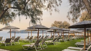 Een rij ligbedden met rieten parasols, een stenen pad en uitzicht op het water.