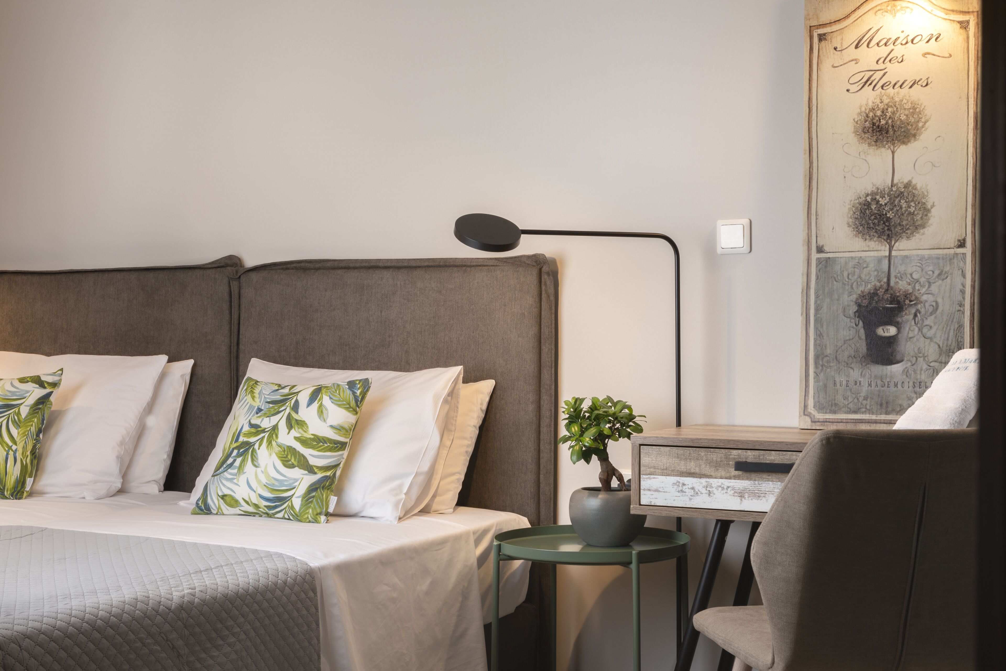 Un dormitorio ordenado con una cama, almohadas, una mesita de noche con una planta y una silla.