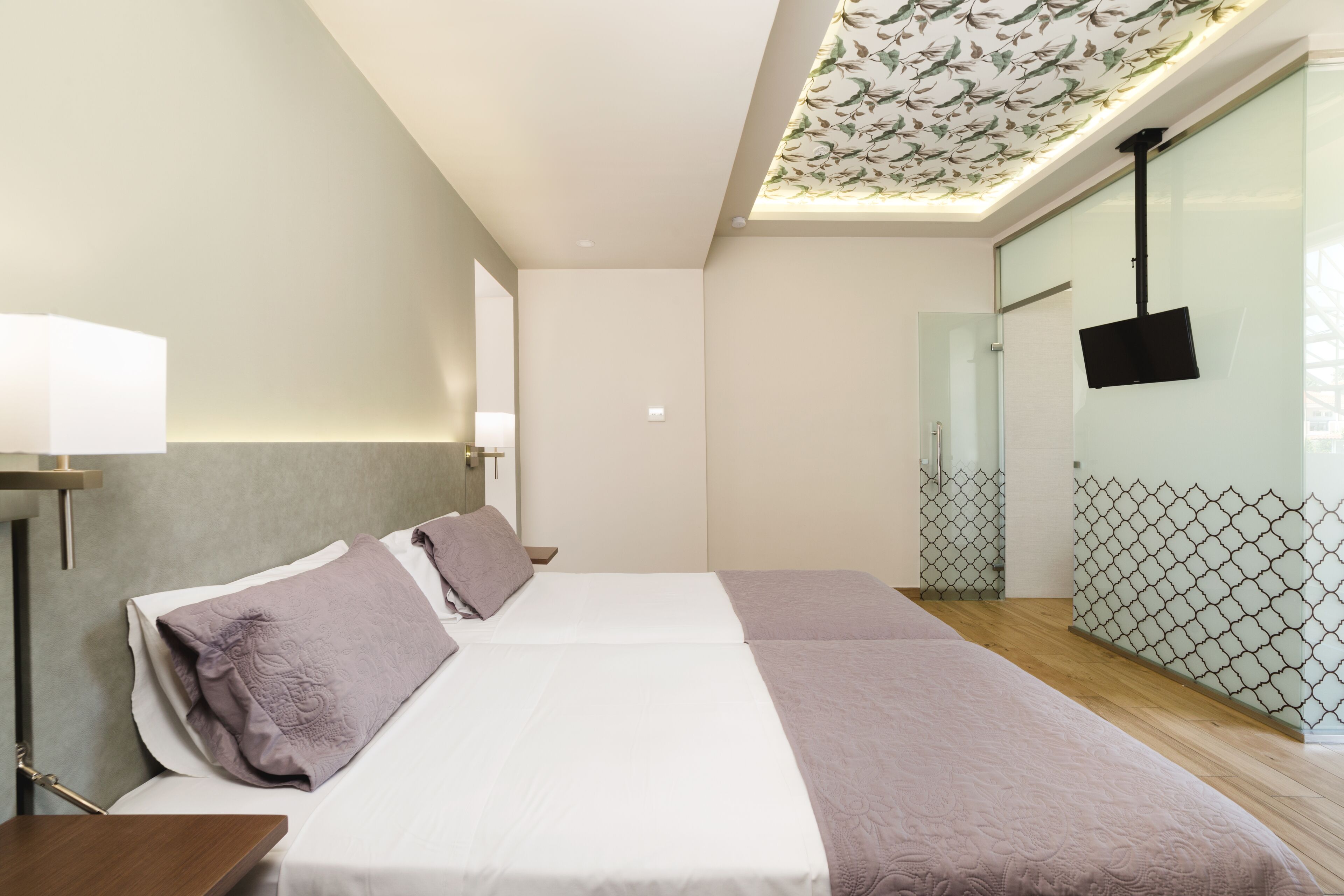 A modern hotel room with a large bed, a bedside table, a flat-screen TV mounted on the wall, and a ceiling with a floral pattern.