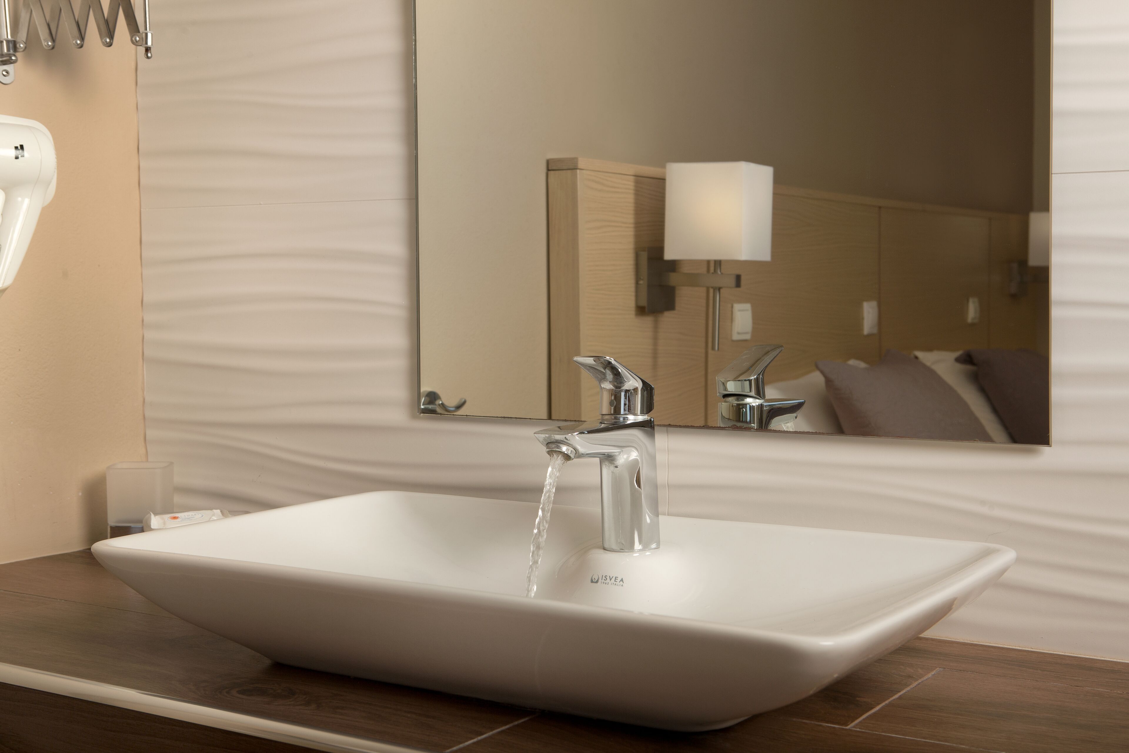 A modern bathroom with a white vessel sink, chrome faucet, and a mirror reflecting a bed with a pillow.