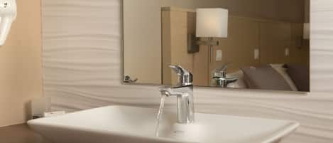 A modern bathroom with a white vessel sink, chrome faucet, and a mirror reflecting a bed with a pillow.
