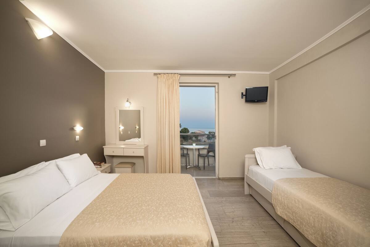 Une chambre d’hôtel avec deux lits, un bureau, une chaise, une télévision et un balcon équipé d’une table et de chaises.