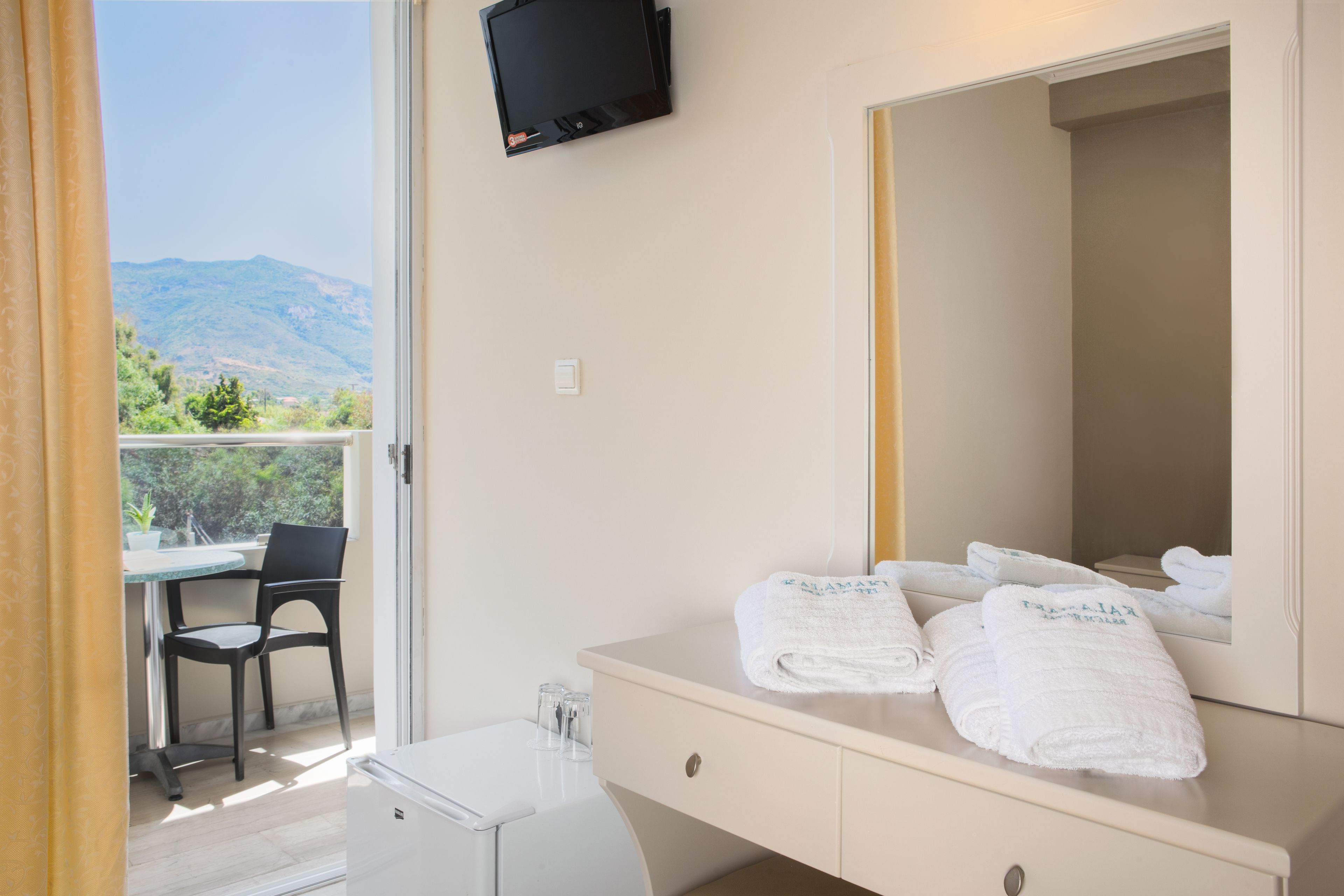 A hotel room with a balcony, a TV mounted on the wall, a white vanity with a mirror, and two towels on the vanity.