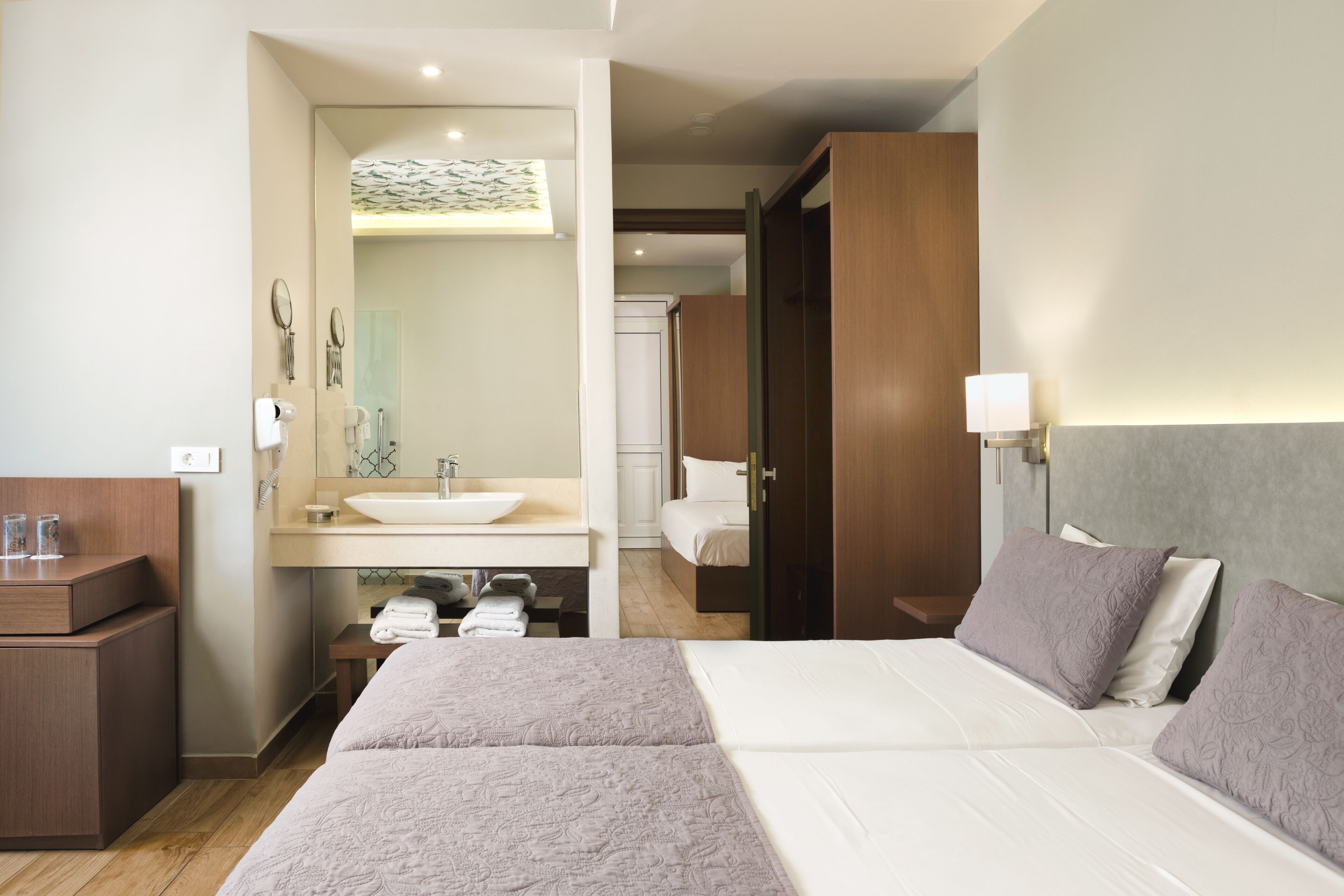 A hotel room with a large bed, a bedside table, a wooden wardrobe, a bathroom with a sink and mirror, and a shower area.