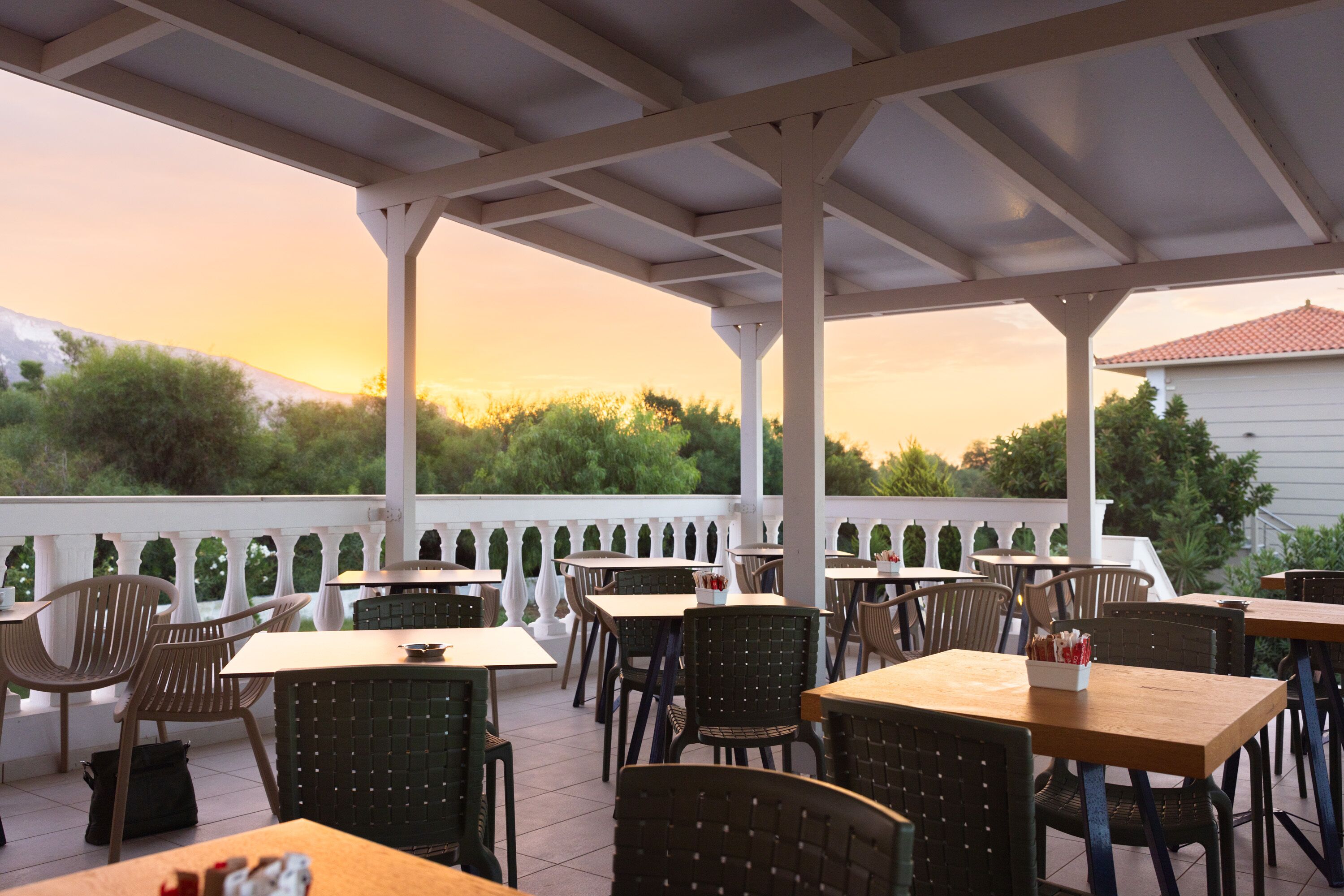 An outdoor patio with tables and chairs, a white railing, and a view of trees and a building in the background.