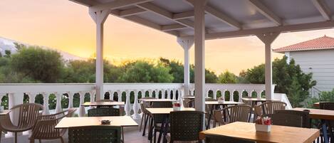 An outdoor patio with tables and chairs, a white railing, and a view of trees and a building in the background.