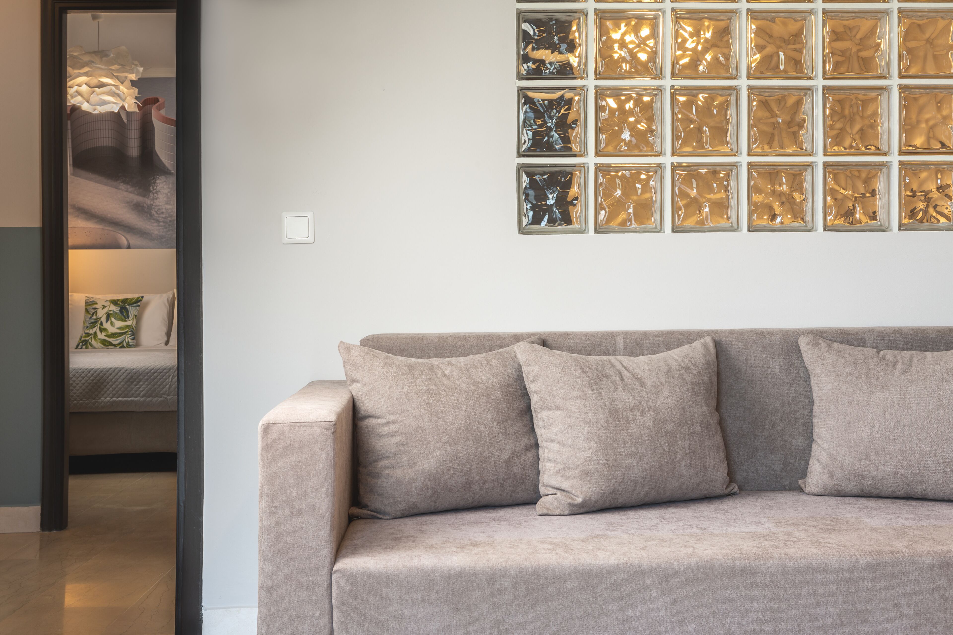 A beige sofa with three cushions, a wall with a decorative glass panel, and a doorway leading to a bed with a patterned pillow.