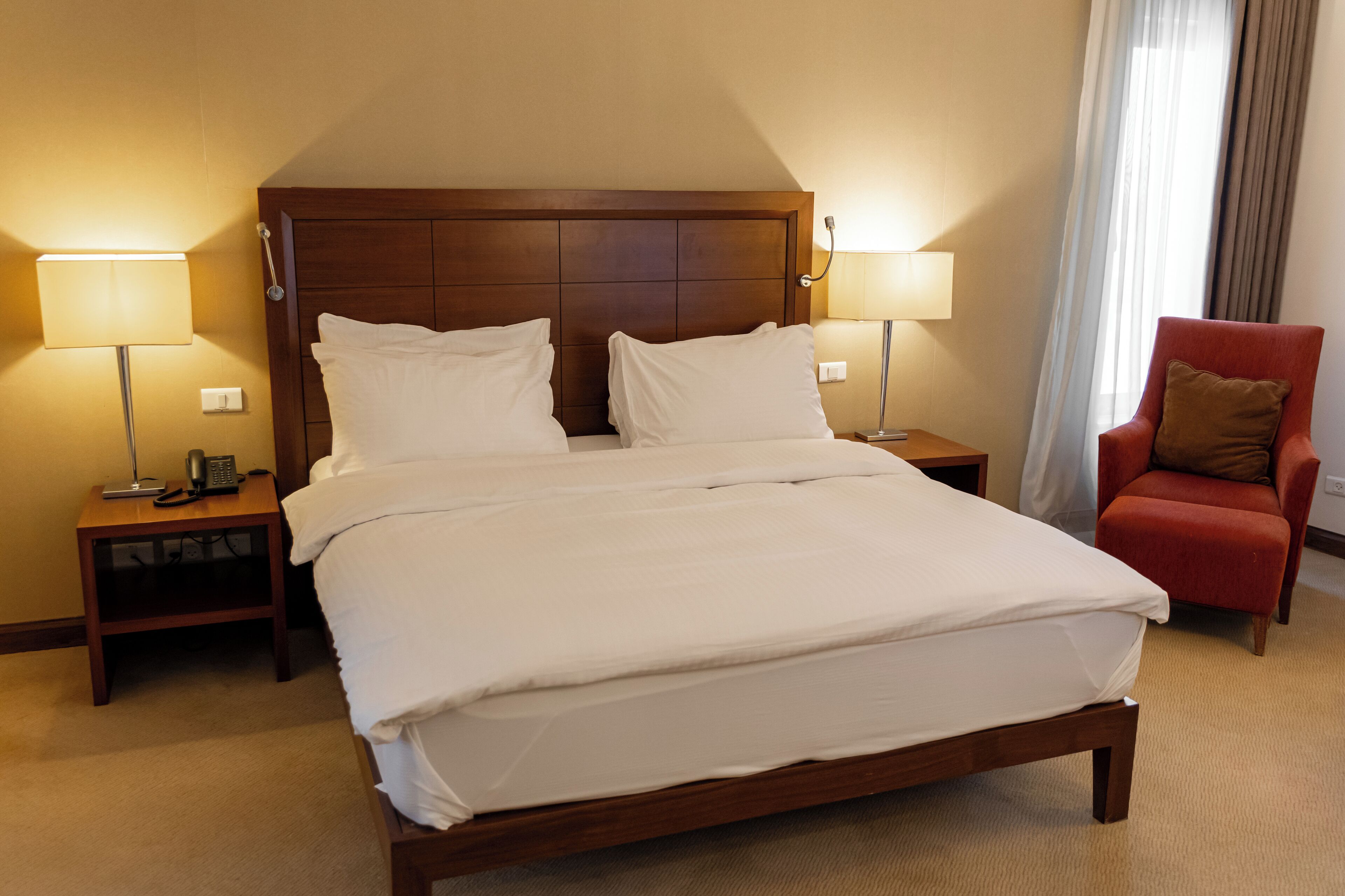 A hotel room with a large bed, two bedside tables with lamps, a red armchair, and a wooden headboard.