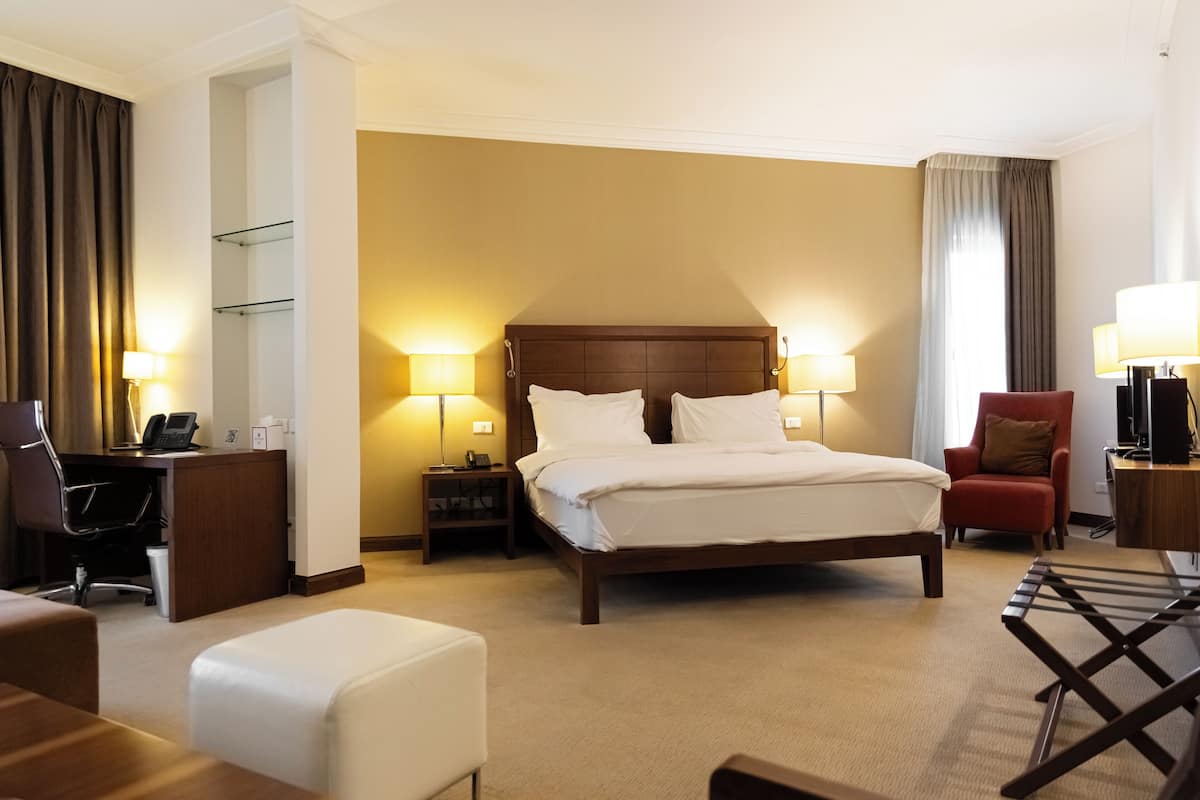 Quarto de hotel com uma cama grande, mesas de cabeceira com luminárias, uma escrivaninha, uma cadeira e uma televisão.
