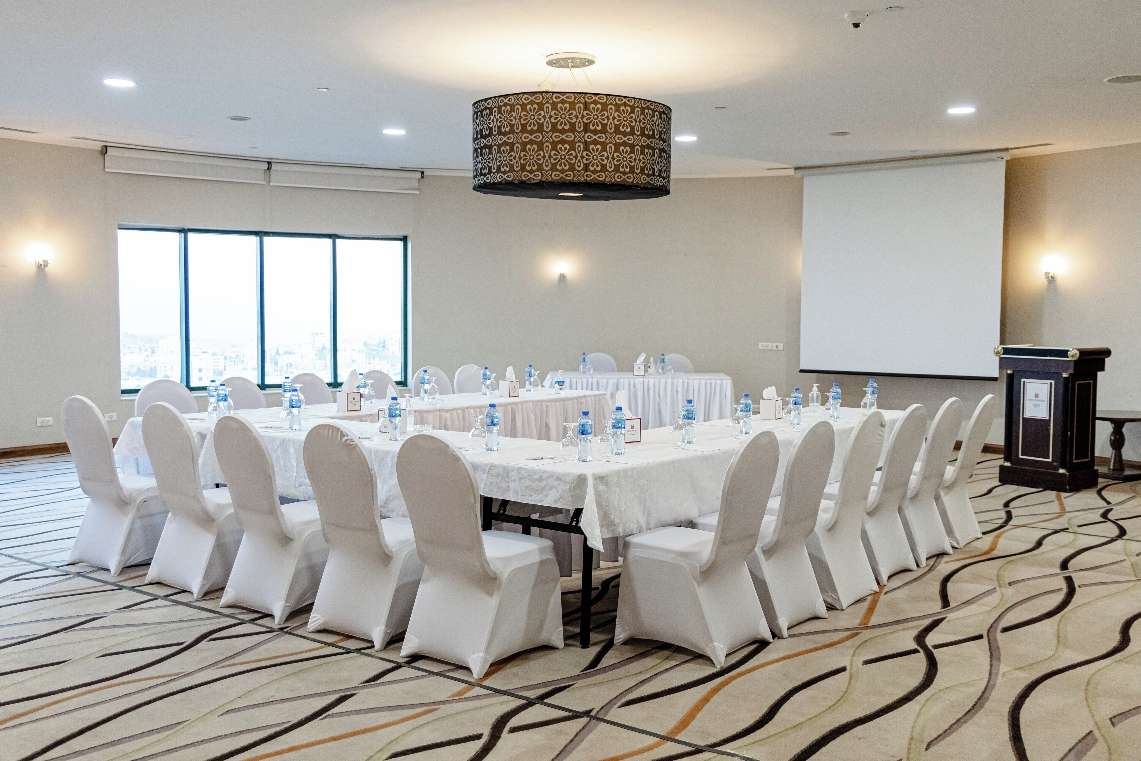 Un salón de conferencias con una larga mesa, sillas blancas, botellas de agua y una pantalla de proyector.