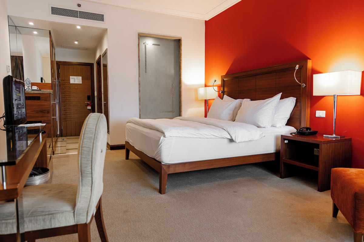 Quarto de hotel com cama, escrivaninha, cadeira e mesa de cabeceira com abajur.