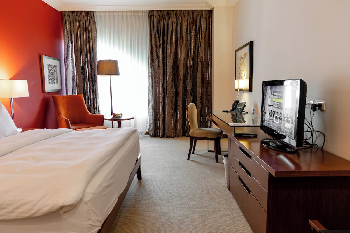 Quarto de hotel com cama, escrivaninha, cadeira, televisão, abajur e cortinas.