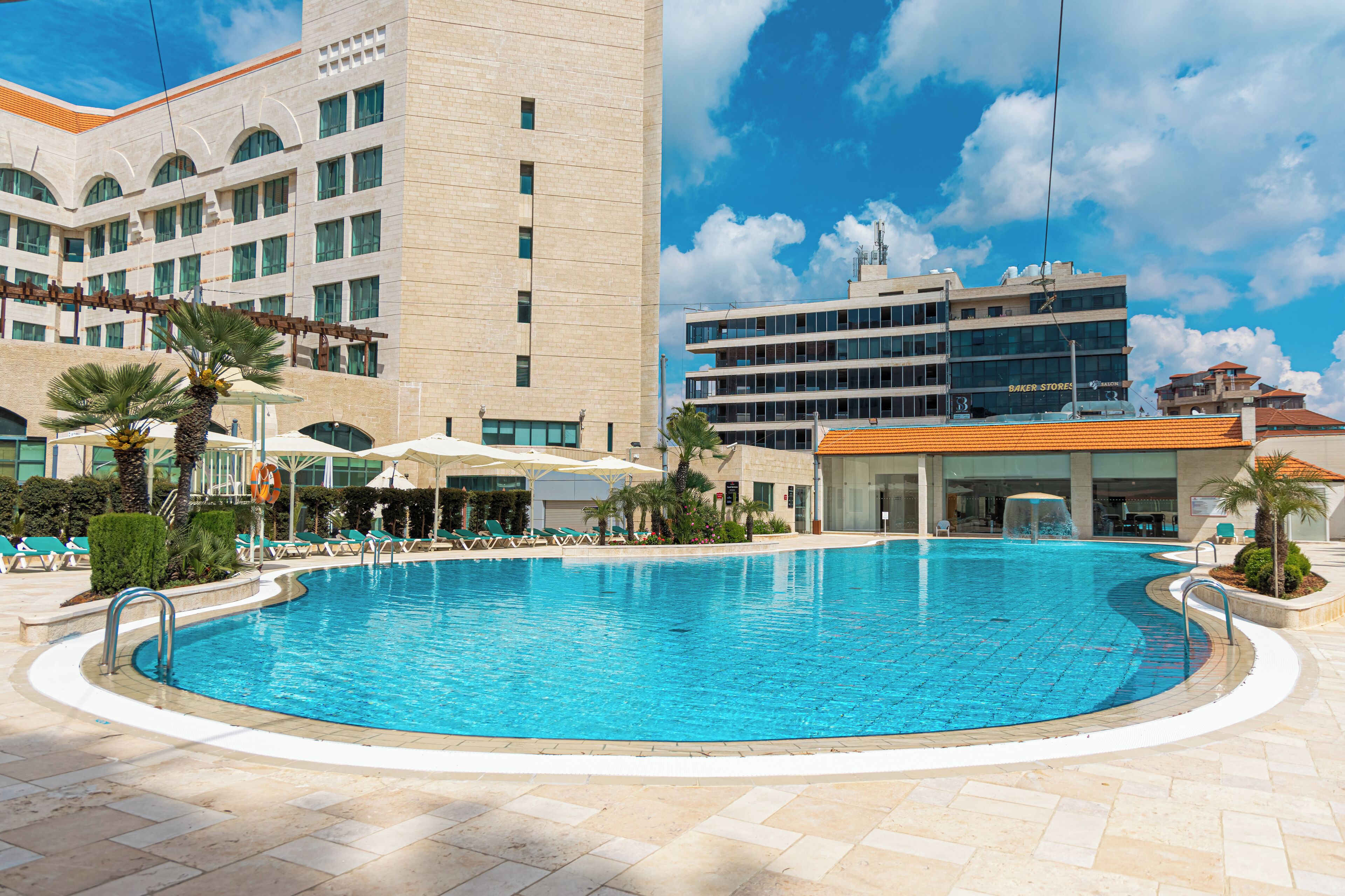 Una piscina con sillones de descanso y sombrillas, rodeada de edificios y palmeras.