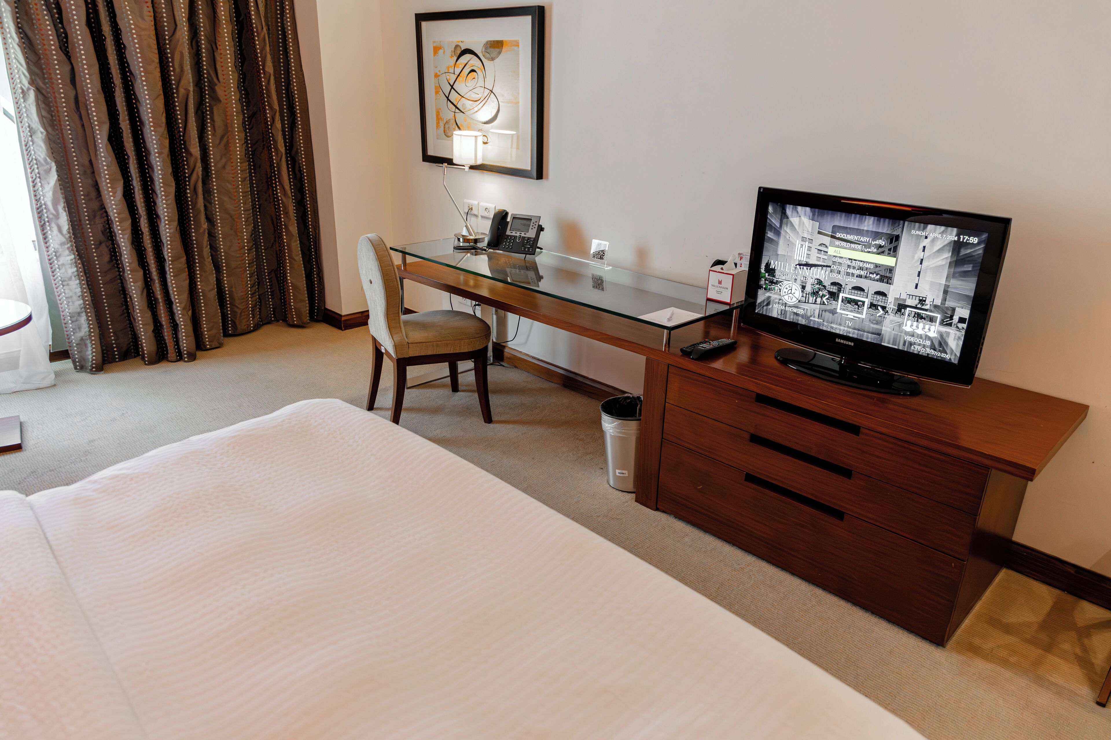 A hotel room with a bed, a desk with a chair, a television, and a trash can.