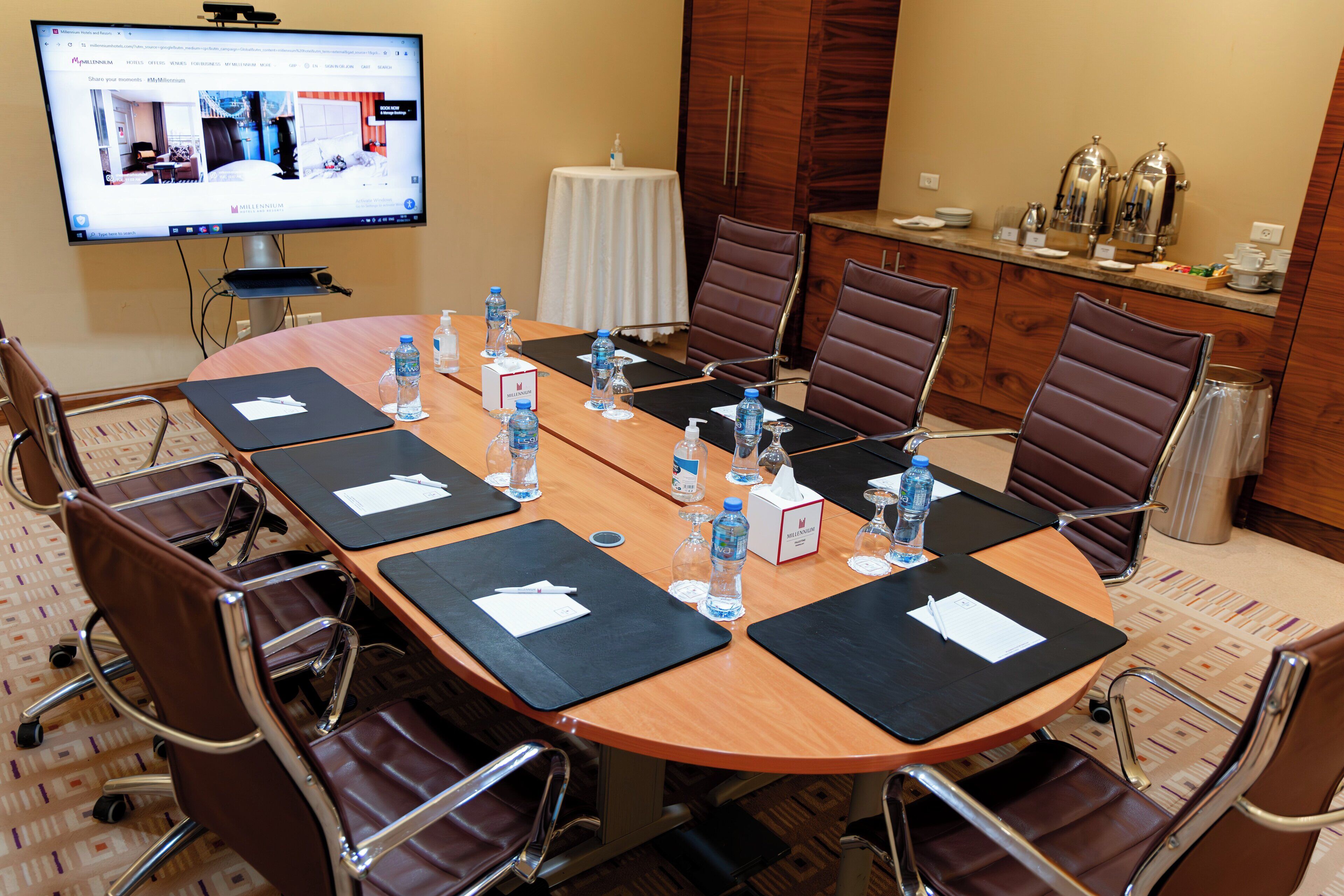 A conference room with a large table, chairs, water bottles, a coffee station, and a television displaying a hotel website.