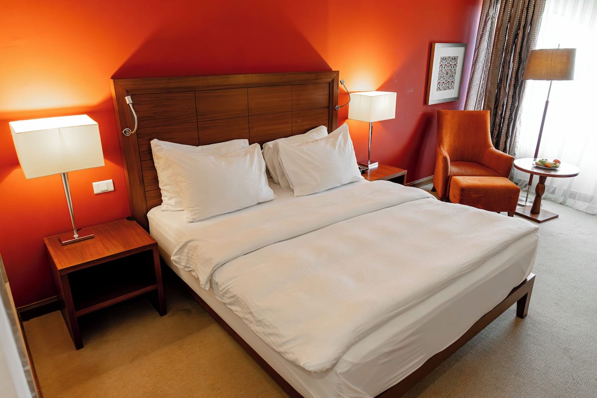 Quarto de hotel com uma cama grande, duas mesas de cabeceira com luminárias, um painel de cabeceira de madeira, uma cadeira e uma pequena mesa com uma cesta de frutas.