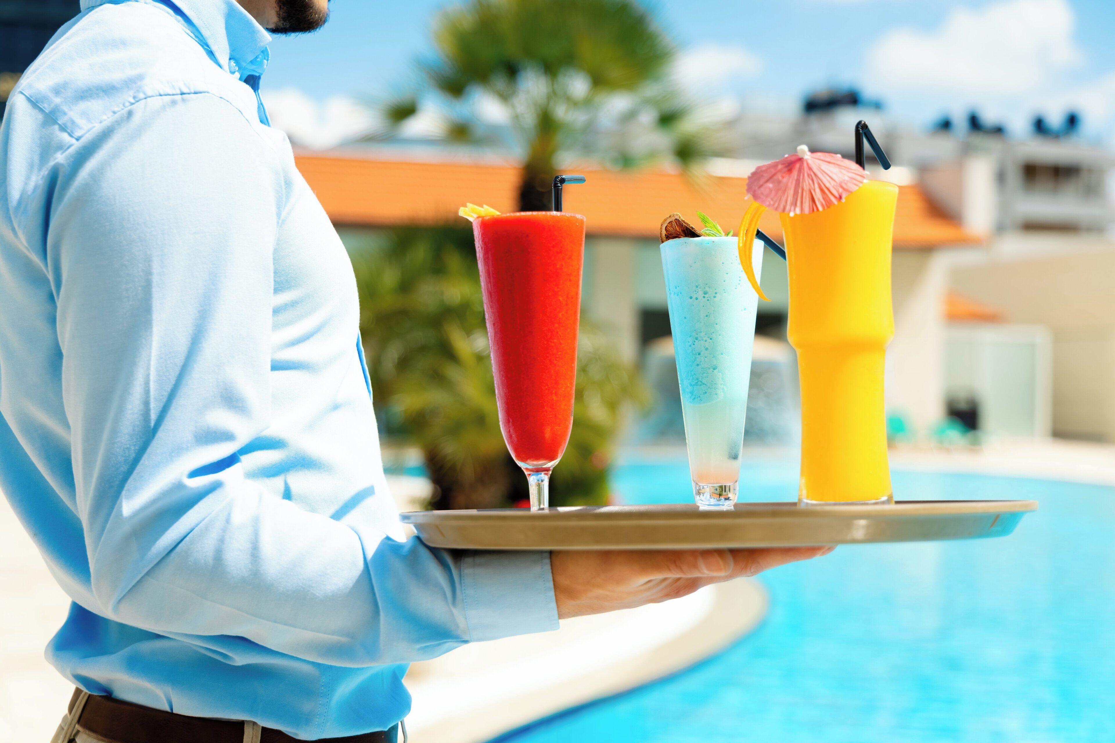 A person holding a tray with three colorful drinks, a pool, and a building with a red roof in the background.