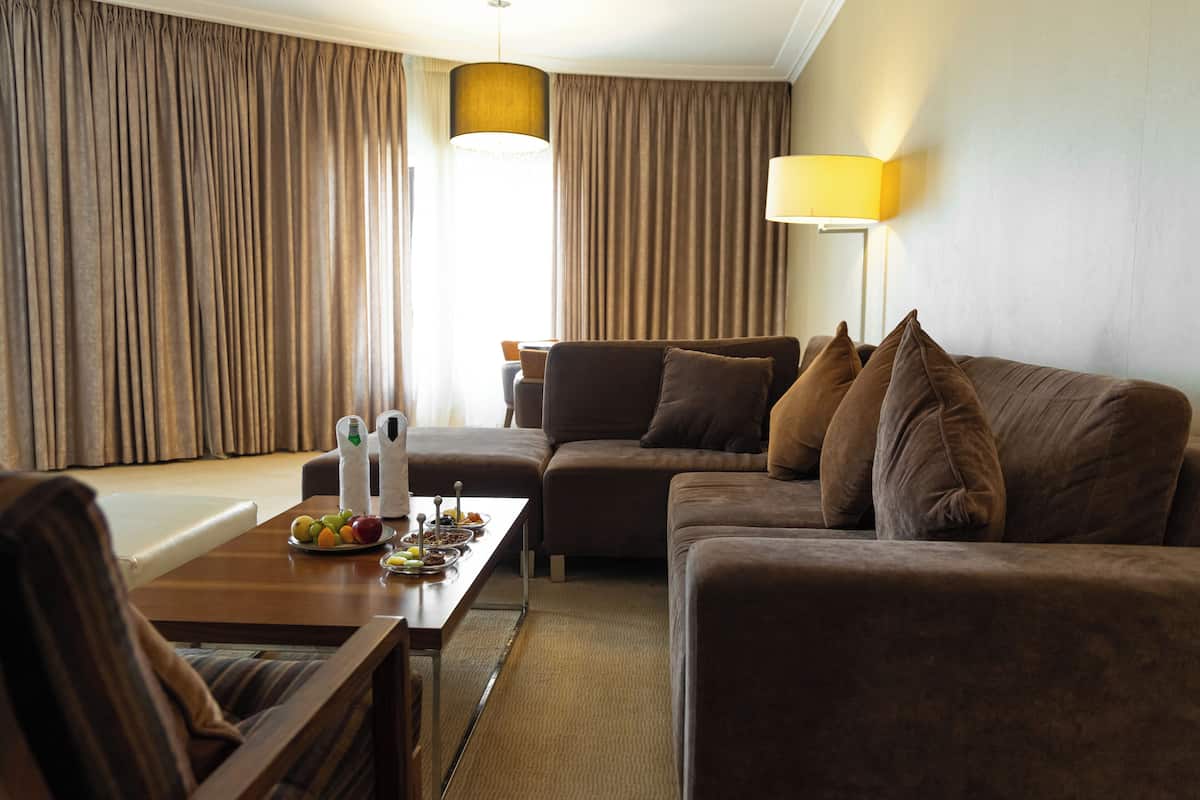 Quarto de hotel com um sofá marrom, uma mesa de centro de madeira com frutas e uma garrafa, uma luminária e cortinas.