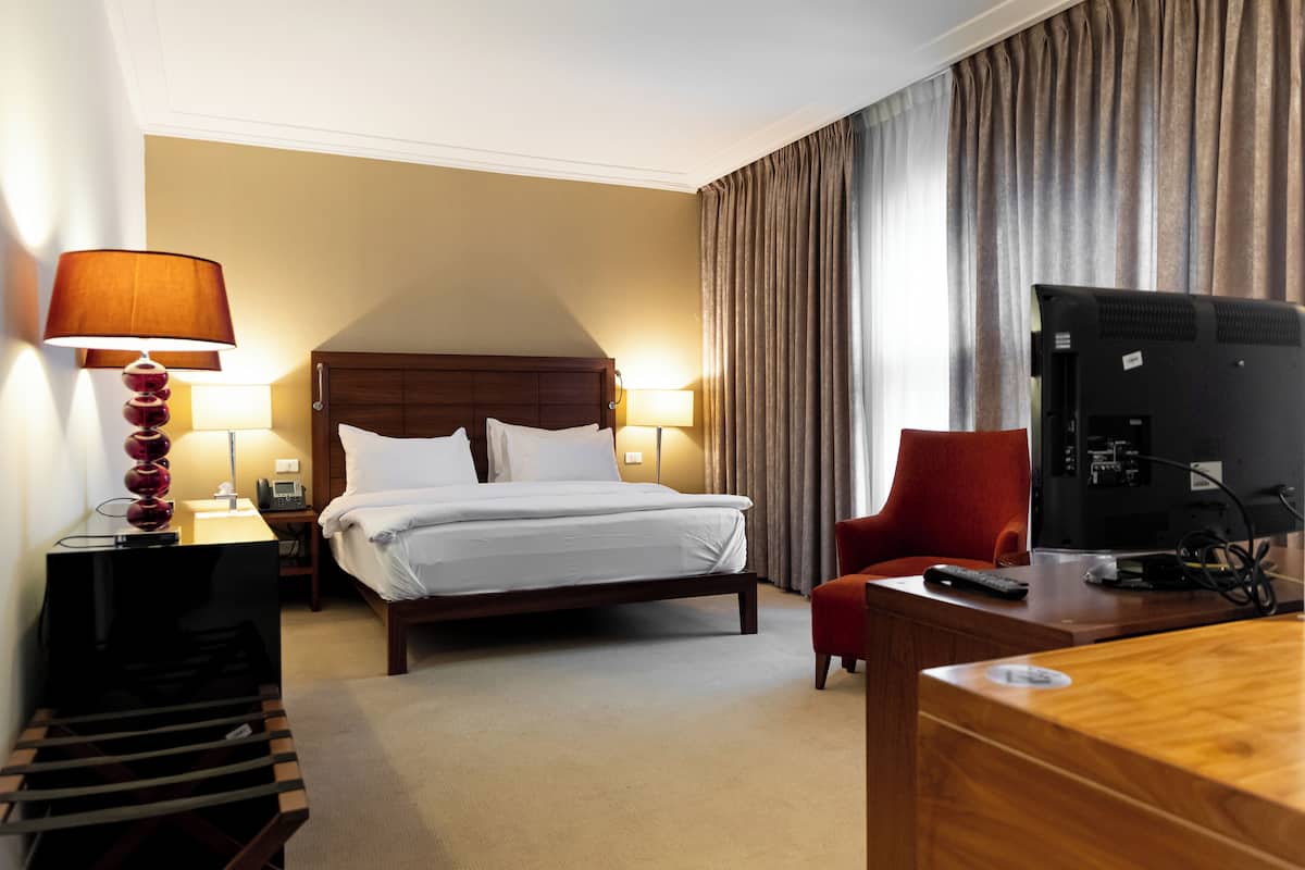 Quarto de hotel com cama, abajures, uma escrivaninha com computador e televisão.