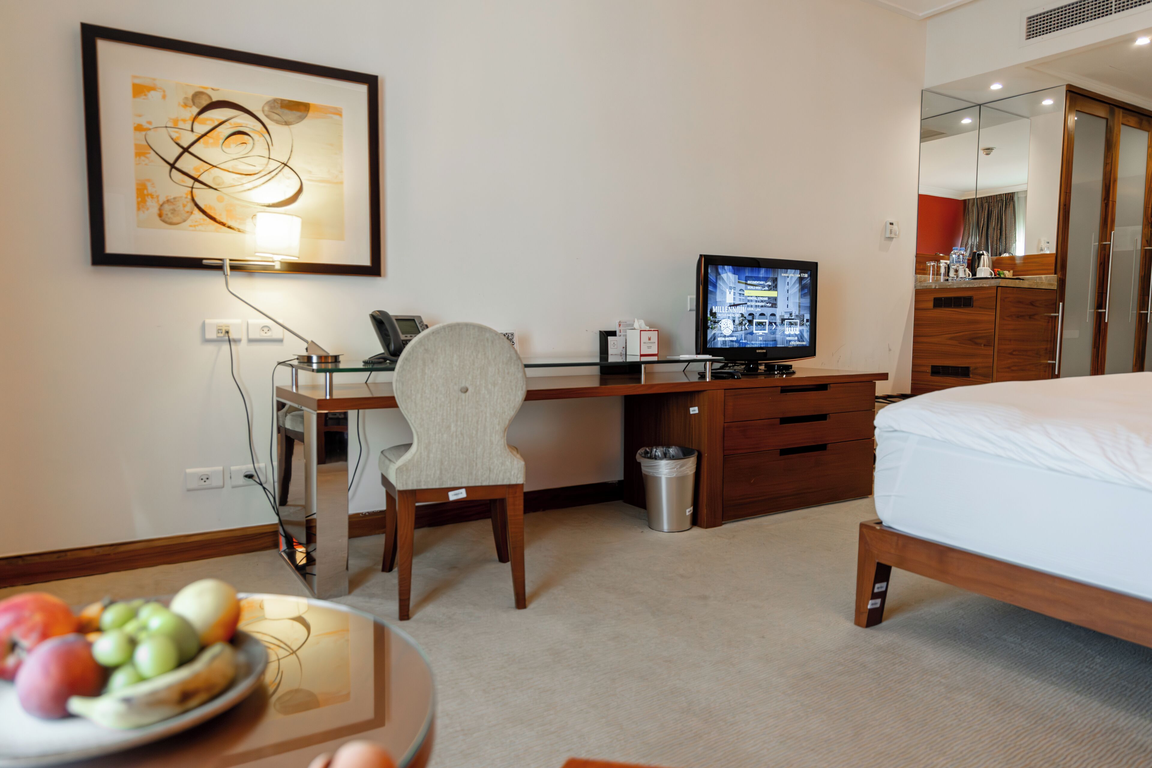 Habitación de hotel con una cama, un escritorio con computadora, una silla, un televisor, un cesto de basura y una obra de arte enmarcada en la pared.